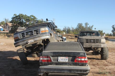 Black 1992 Ford Bronco lifted high, front wheel on Volvo sedan, with another Ford truck behind