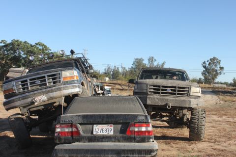 Two lifted Ford trucks, one on top of a Volvo sedan, and another Ford truck in front