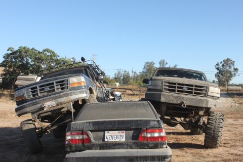 Two lifted Ford trucks, one on each side of a black Volvo sedan, on a dirt lot