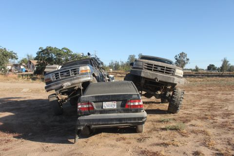 Two lifted Ford trucks are positioned around a black sedan. The truck on the left is a dark color, the truck o