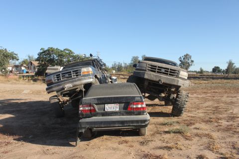 Two lifted Ford trucks are positioned over a sedan in a dirt lot. One truck is on the left, the other on the r
