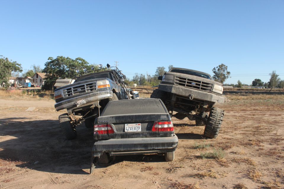 Two lifted Ford trucks are positioned over a sedan in a dirt lot. One truck is on the left, the other on the right