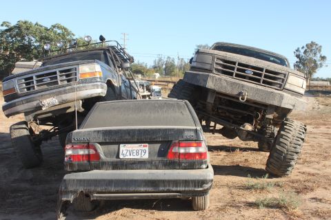 Two lifted Ford trucks and a Volvo sedan are parked in a dirt lot