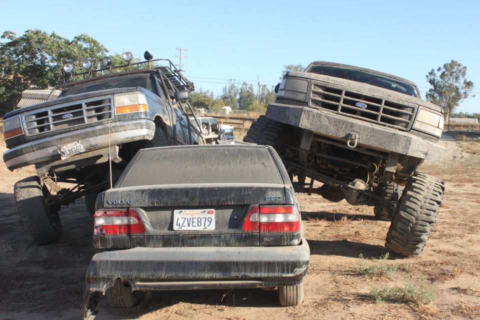 Two lifted Ford trucks and a Volvo sedan are parked in a dirt lot