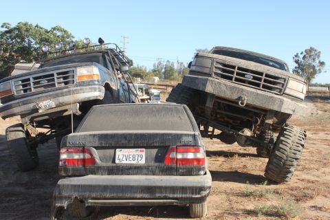 Two lifted Ford trucks tower over a Volvo sedan in a dusty outdoor scene