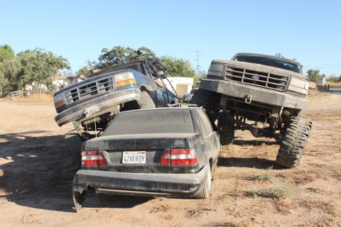 Two lifted Ford trucks are stacked on top of a black sedan. The sedan has a license plate that reads 4ZVE879
