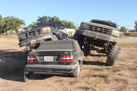 1992 Ford Bronco, dark blue, lifted with large tires, on a sedan. Another lifted Ford truck is nearby