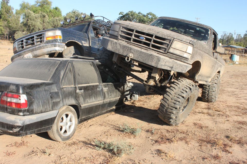 Two heavily modified Ford trucks are stacked on top of a black sedan in a dirt lot