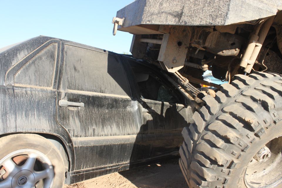 Black sedan with a crushed side, partially obscured by a large off-road vehicle's tire and chassis