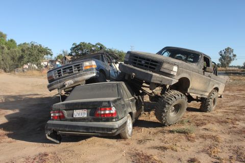 Two lifted trucks are stacked on top of a black sedan in a dirt lot