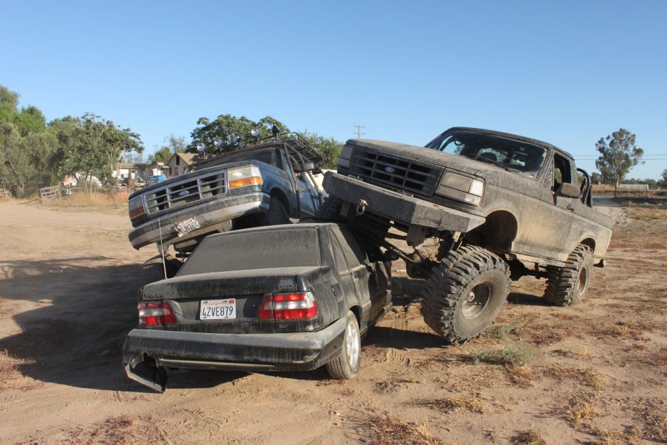 Two lifted trucks are stacked on top of a black sedan in a dirt lot