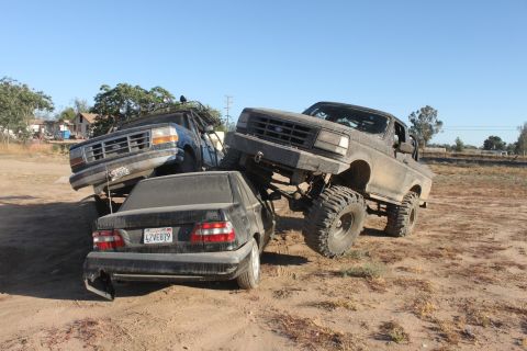 Two lifted Ford trucks on a dirt lot, one crushing a sedan