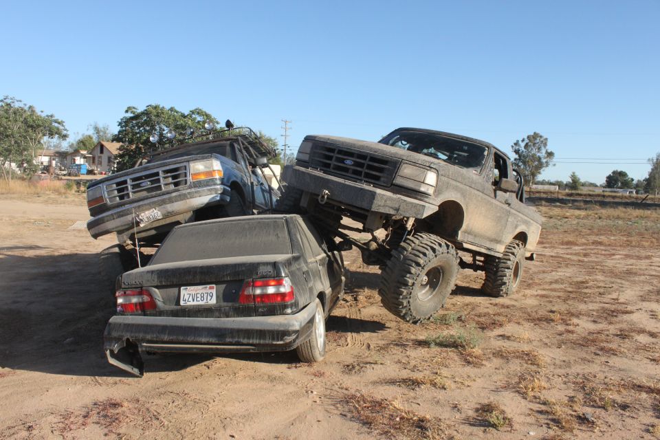 Two lifted Ford trucks on a dirt lot, one crushing a sedan