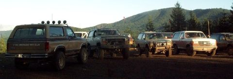 1982 Ford Bronco - Several SUVs are parked on a dirt lot with trees and hills in the background.