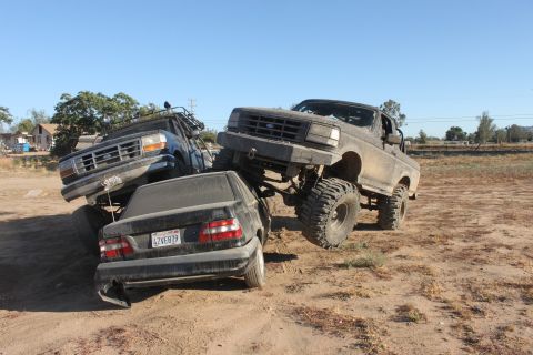 Two lifted Ford trucks are crushing a black sedan in a dirt lot. The front truck is a dark color, the rear tru