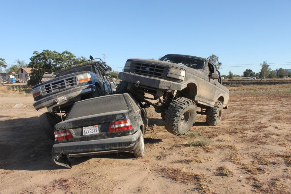 Two lifted Ford trucks are crushing a black sedan in a dirt lot. The front truck is a dark color, the rear truck is blue. The sedan's license plate is visible