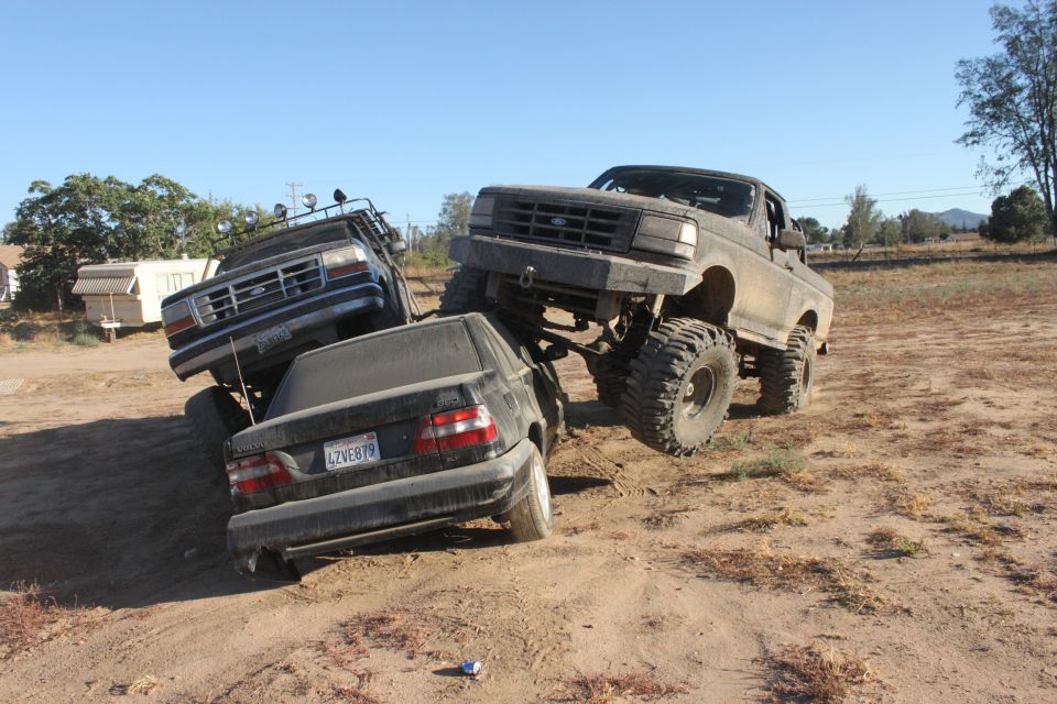 Two lifted trucks are on top of a sedan in a dirt lot. One truck is a Ford Bronco