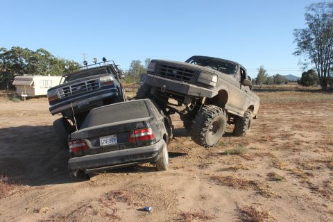 Mud-covered 1992 Ford Bronco lifted over a dark sedan and another pickup truck on a dirt lot