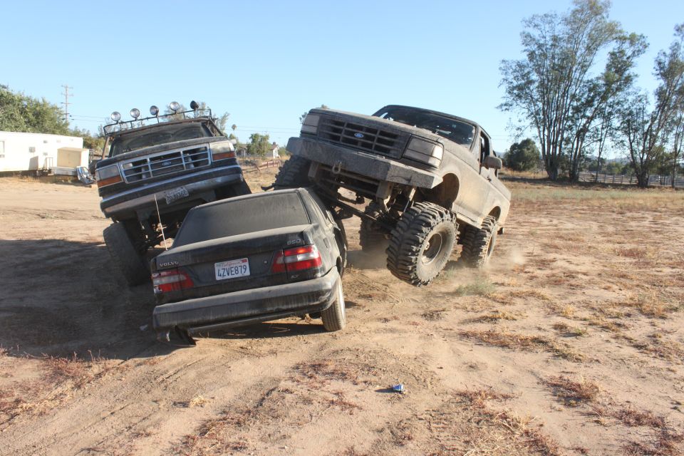 Two lifted trucks are crushing a black sedan in a dirt lot. One truck is on the right, the other on the left