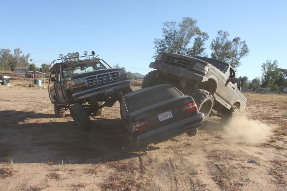 Two lifted Ford trucks crushing a dark sedan on a dirt lot