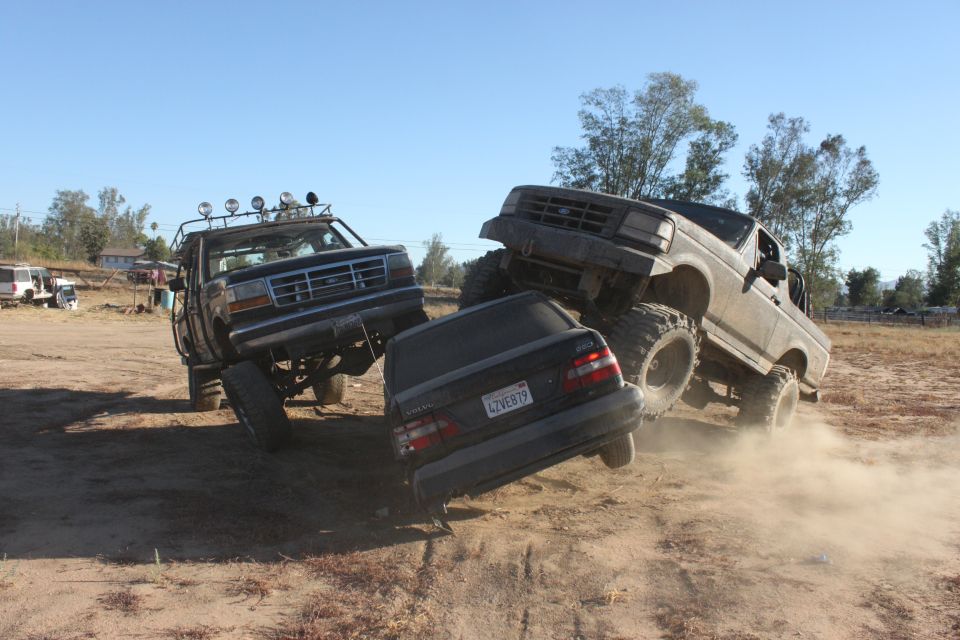 Two monster trucks are crushing a black sedan. One truck is on the left, the other is on the right, driving over the car