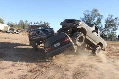 1992 Ford Bronco lifted and driving over a sedan, with another lifted truck nearby