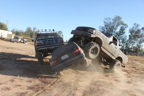 Two lifted trucks crushing a car on a dirt hill. 1992 Ford Bronco visible