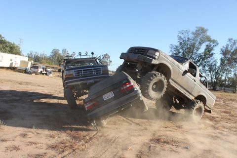 1992 Ford Bronco and another lifted truck crushing a sedan on a dirt lot