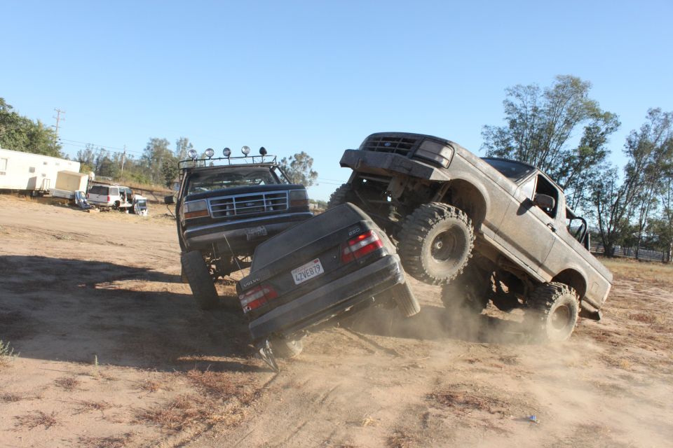 1992 Ford Bronco and another lifted truck crushing a sedan on a dirt lot