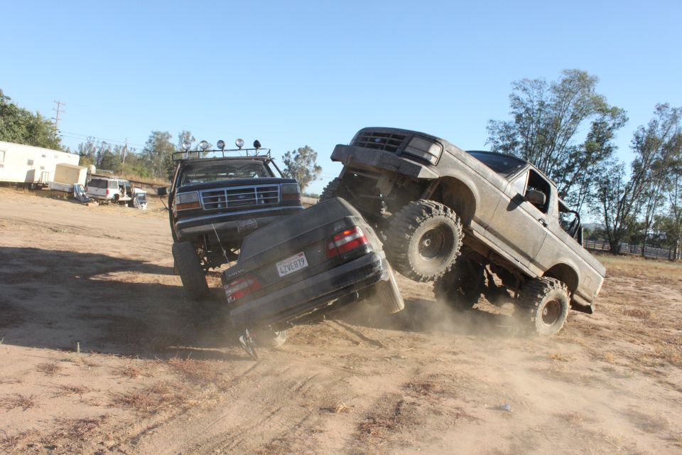 Two lifted trucks are crushing a car on a dirt hill. One truck is on the right, the other on the left