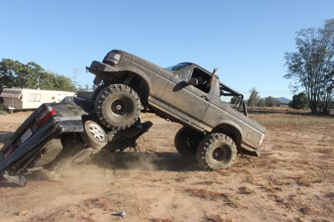 Mud-covered 1992 Ford Bronco on top of a crushed black sedan, with a person giving a peace sign from the drive