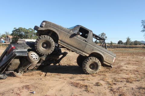 Muddy 1992 Ford Bronco on top of a crushed car, off-road scene