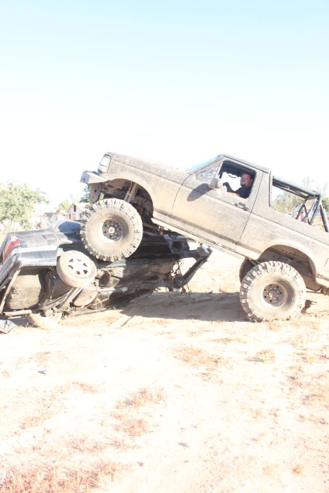 Mud-covered 1992 Ford Bronco on top of a crushed vehicle, driver visible inside
