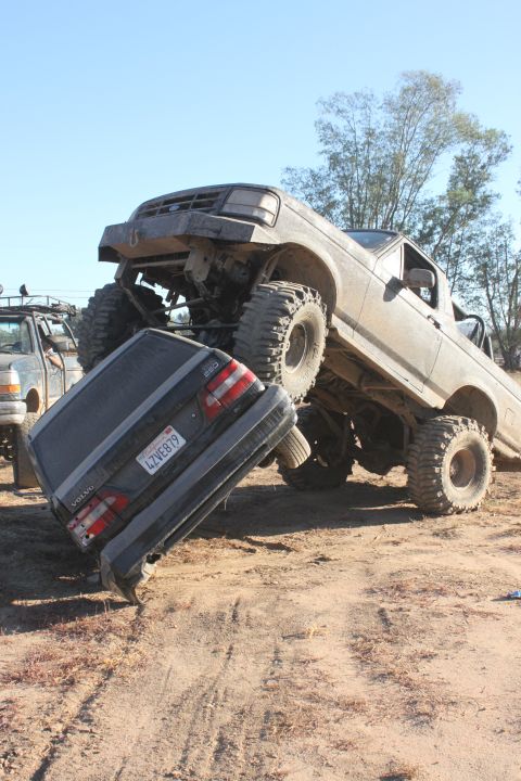 1992 Ford Bronco, lifted and modified, crushing a dark Volvo sedan on a dirt track