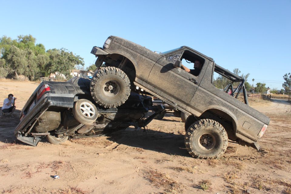 Mud-covered 1992 Ford Bronco on top of an overturned black car, driver visible inside the Bronco