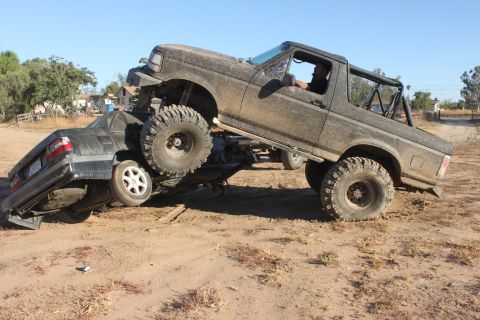 Muddy 1992 Ford Bronco on top of a crushed car, driver visible