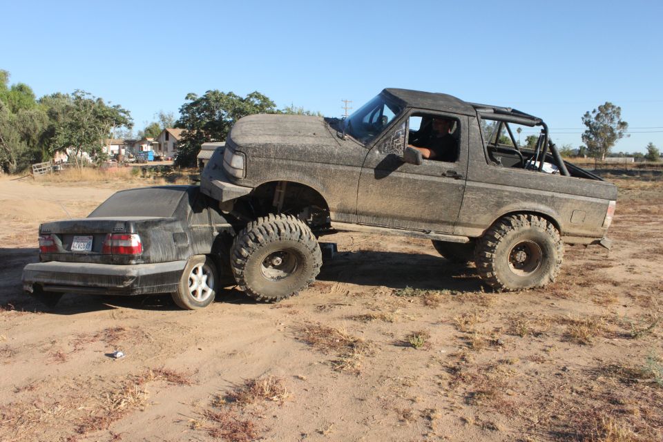 Mud-covered 1992 Ford Bronco on top of a black sedan, front right tire of Bronco on sedan's hood