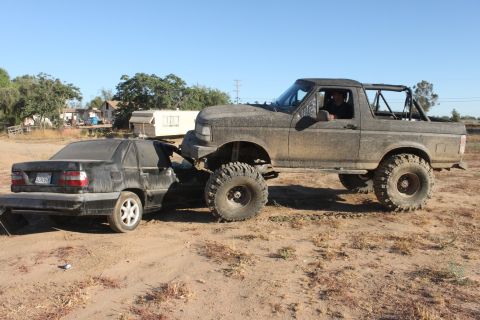 Muddy 1992 Ford Bronco pushing a black sedan on a dirt lot