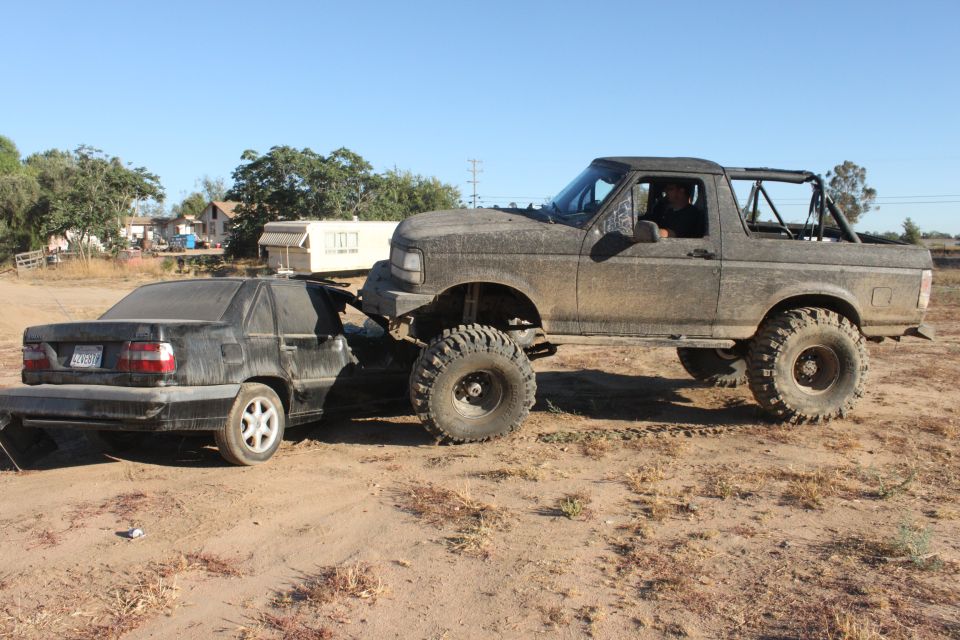 Muddy 1992 Ford Bronco pushing a black sedan on a dirt lot
