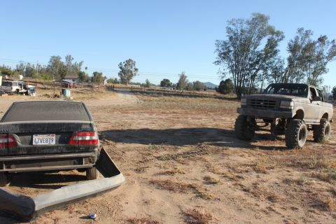 Lifted pickup truck parked in a dry, dusty lot next to a black sedan with its bumper removed. Other vehicles a