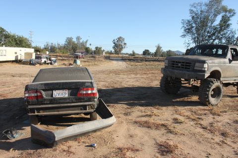 Lifted Ford truck parked next to a Volvo sedan with its bumper detached in a dusty lot