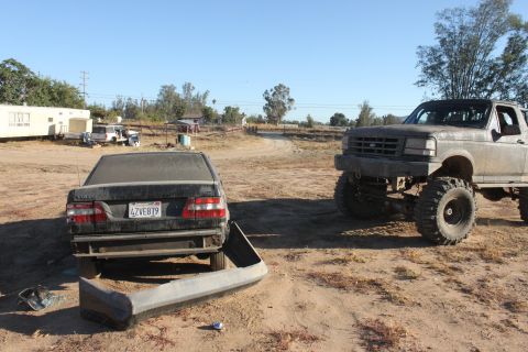 Dusty black sedan with detached bumper and lifted grey truck in a dry, outdoor lot