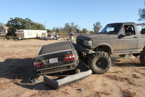 Muddy 1992 Ford Bronco crushing a black sedan. License plate LZVE879 is visible on the sedan