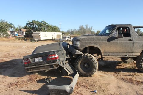 1992 Ford Bronco with large tires crushing a black sedan. The sedan's rear bumper is detached