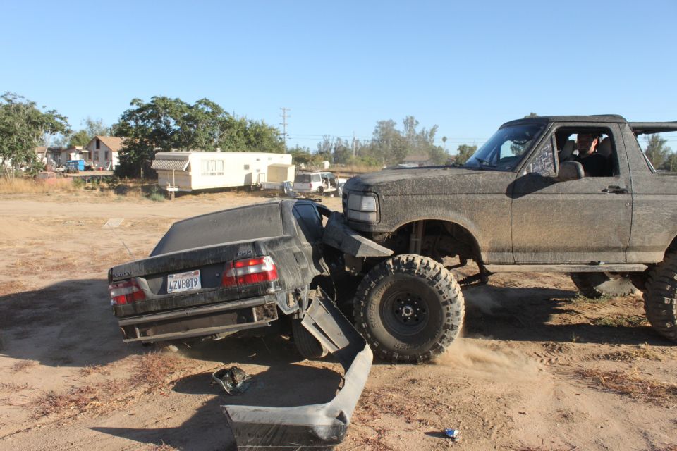 1992 Ford Bronco with large tires crushing a black sedan. The sedan's rear bumper is detached