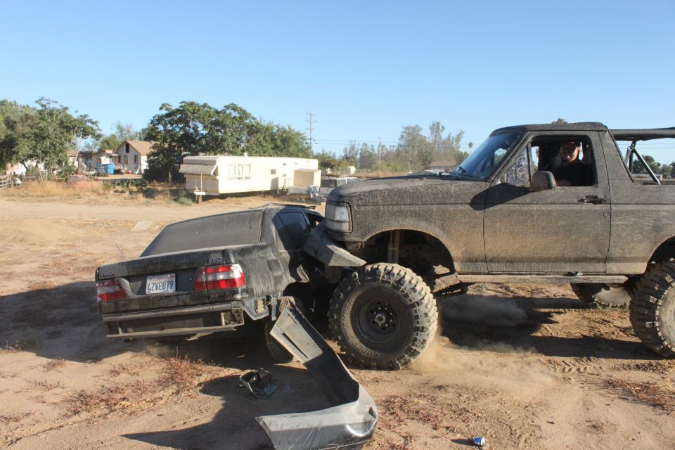 Big lifted truck is crushing a black sedan in a dirt lot. The sedan's bumper is detached