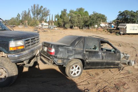 Ford truck is pushing a wrecked black sedan with broken windows in a dirt lot