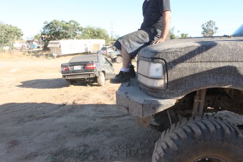 Person sits on front bumper of large truck; sedan in background