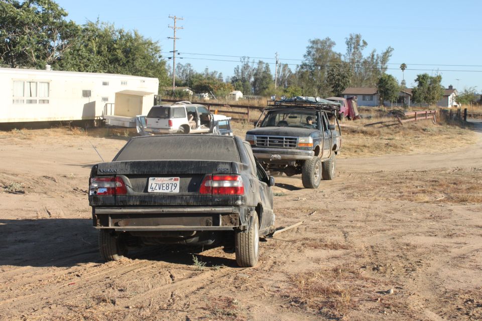 1992 Ford Bronco with roof rack and lifted suspension, followed by a damaged Volvo sedan on a dirt road
