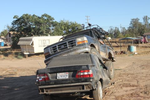 Ford truck on top of a Volvo sedan, parked in a dirt lot with a trailer and houses in background